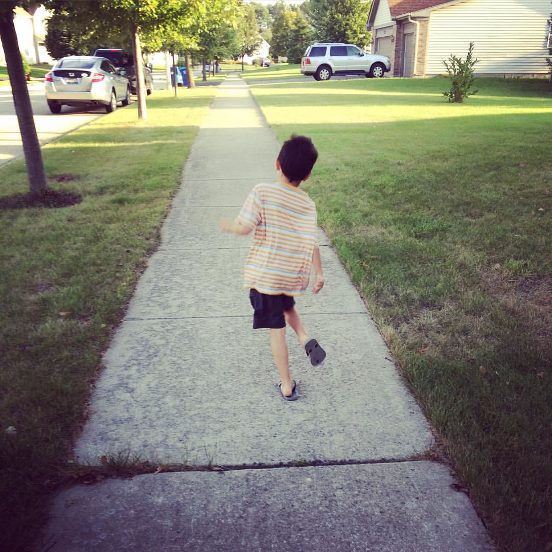 A young boy wearing an orange shirt, shorts, and flip flops running down a sidewalk in a residential neighborhood
